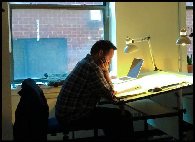 Vinson Milligan concentrating on work at a desk by the window with laptop and lamps at University of the Arts Museum Exhibiition Planning and Design studio