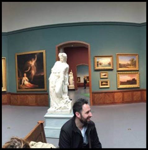 Vinson Milligan sitting down on a bench in front of a statue surrounded by paintings at the Academy of Fine Arts gallery in Philadelphia